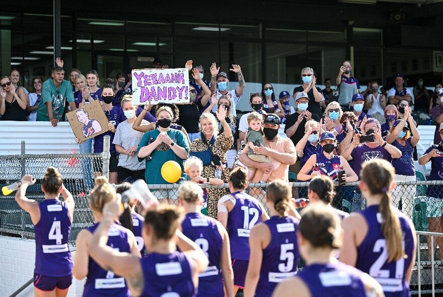 An AFLW team wearing Fremantle jumpers leaving a sports ground, while the fans in the crowd in the covered pavillion clap and cheer them.