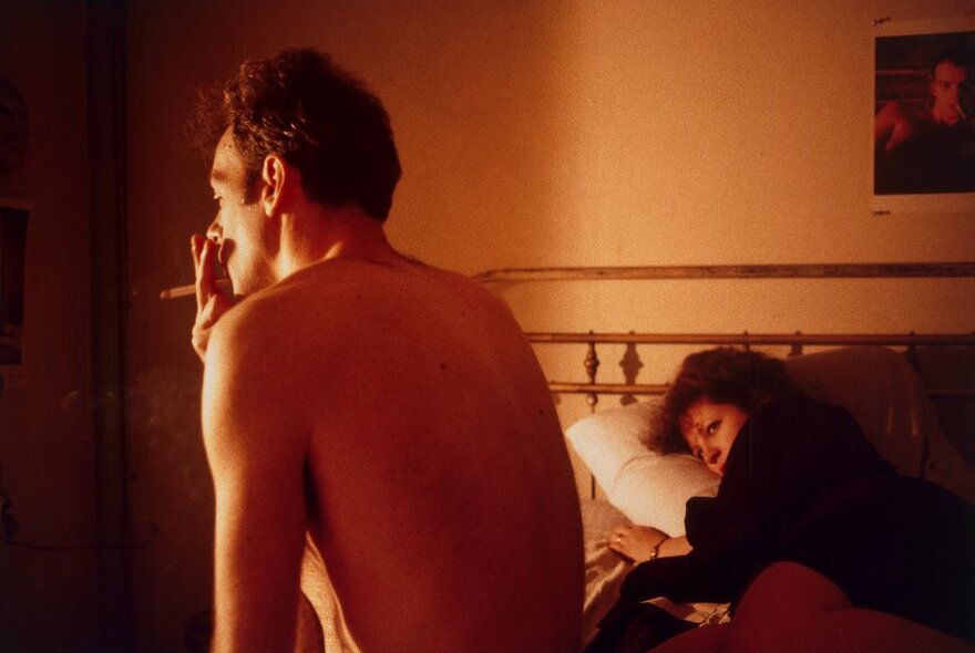 Man sitting on the edge of a bed with a cigarette, while a woman looks on.