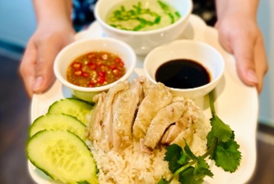 Pair of hands holding a large plate of poached chicken over rice and salad with bowls of sauces of the side.