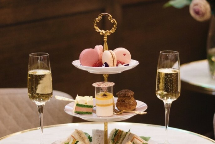 Three-tiered cake dish with cakes, biscuits and sandwiches with two glasses of champagne either side.