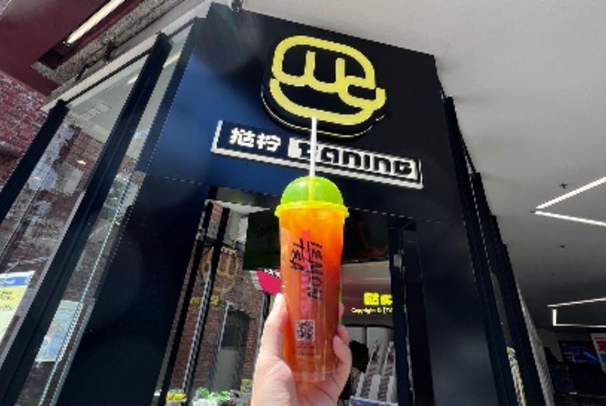 Hand holding a plastic takeaway cup of iced lemon tea in front of the entrance to the Taning Lemontea shop in Melbourne's Chinatown