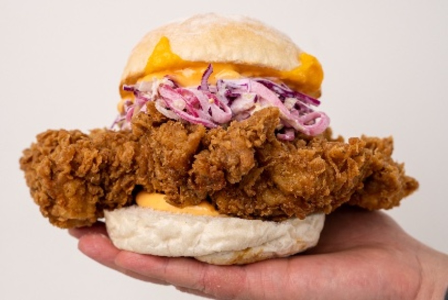 An enormous piece of fried chicken inside a smaller hamburger bun, with cheese and coleslaw.