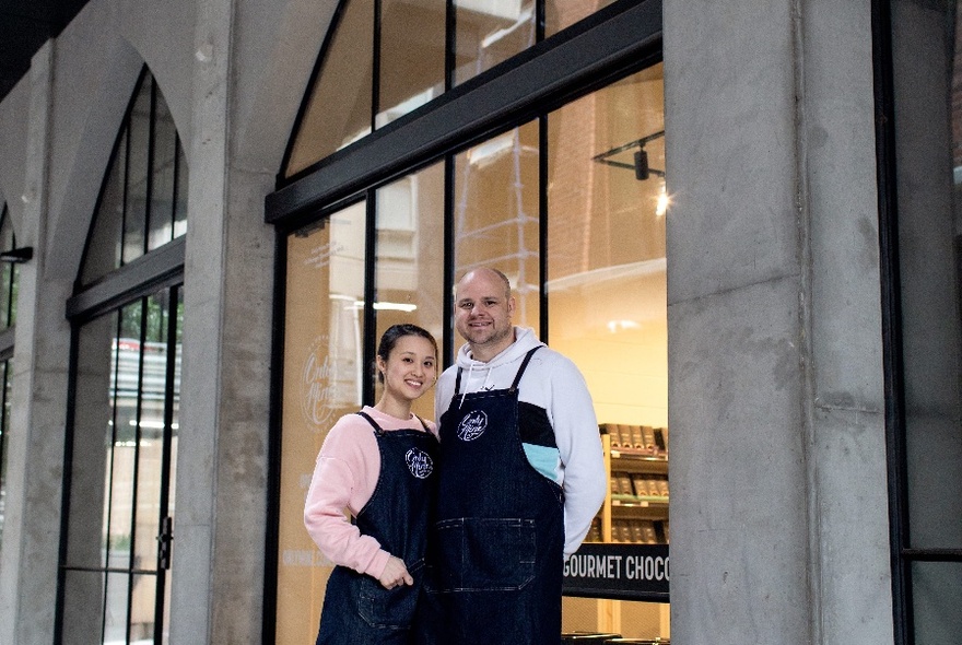 A man and an woman pose in front of a large window of a grey stone shop with an arm around each other. 