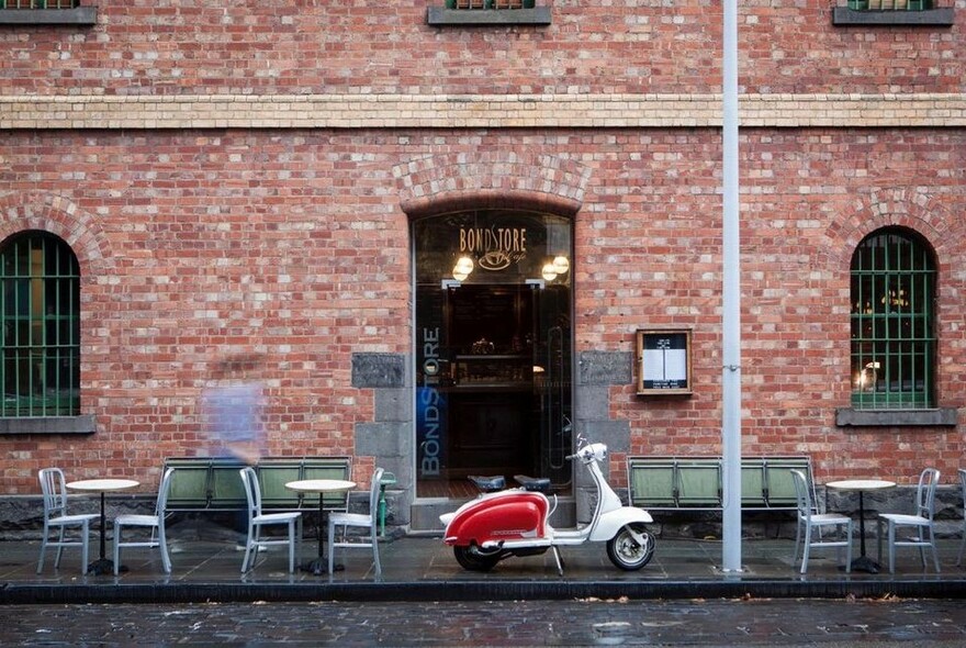 Red brick cafe with small tables and chairs outside and a red motor scooter.