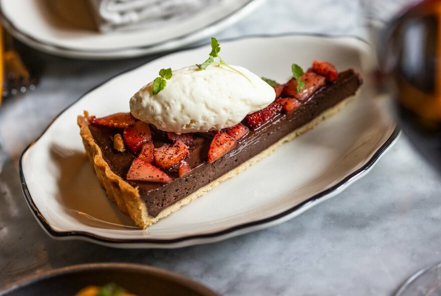 A slice of chocolate tart, garnished with cream and strawberries, resting on an oval plate on a table in a restaurant.