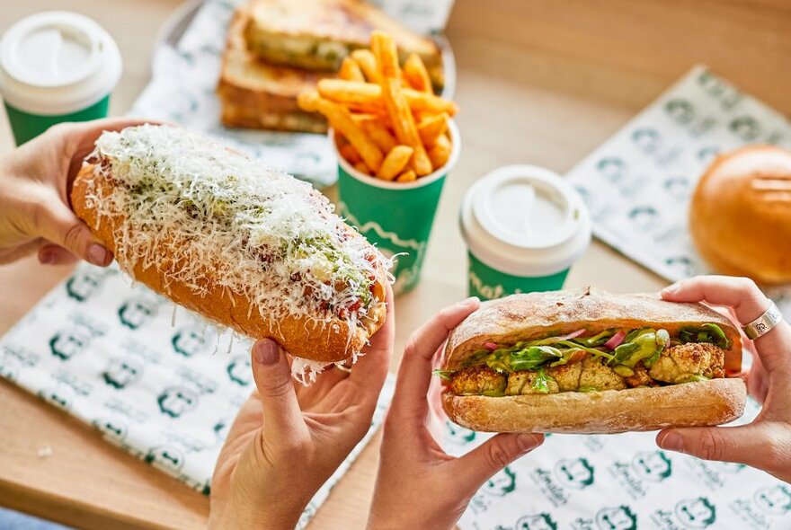 Two pairs of hands holding up sandwiches in long bread rolls, one roll covered in grated cheese, hot chips and takeaway coffee cups in the background.