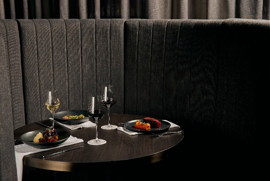 Sleek booth and curved banquette seating in dark charcoal tones, showing a table set with plates of food and three glasses of wine.
