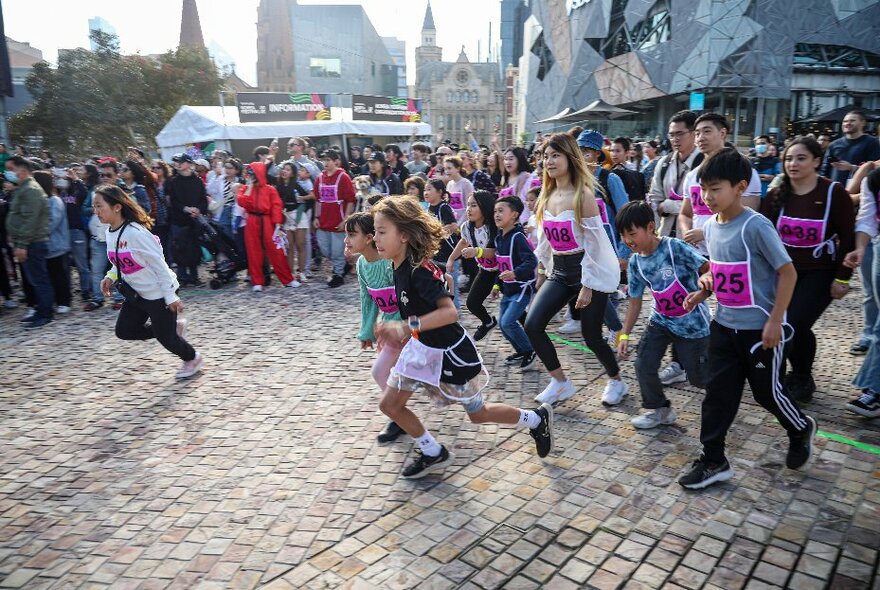 A large group of children of varying ages, wearing racing style numbered bibs on their chests, running around in Fed Square plaza as part of the Korea Festival.