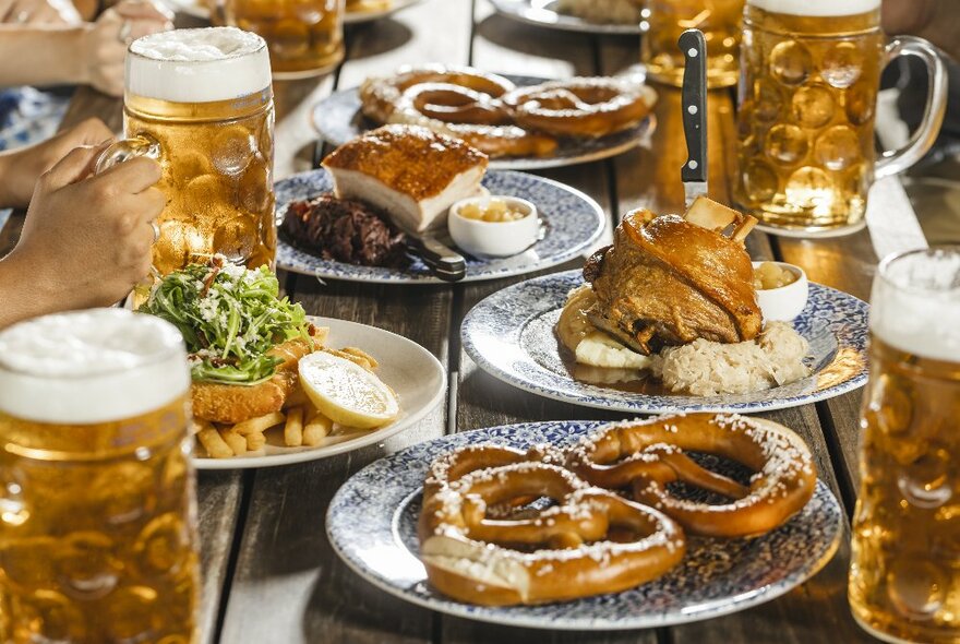 A wooden table filled with a spread of Bavarian food including plates of large pretzels and ham hock, and steins of beer. 