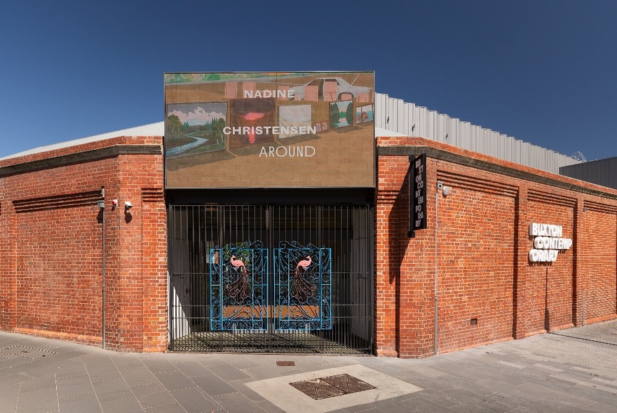 Exterior of redbrick, low-rise Buxton Contemporary building with a small billboard over the gated entrance announcing the current exhibition on display.