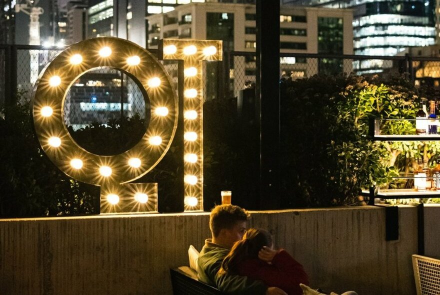 People seated in a rooftop bar with illuminated QT signage, city buildings in the background.