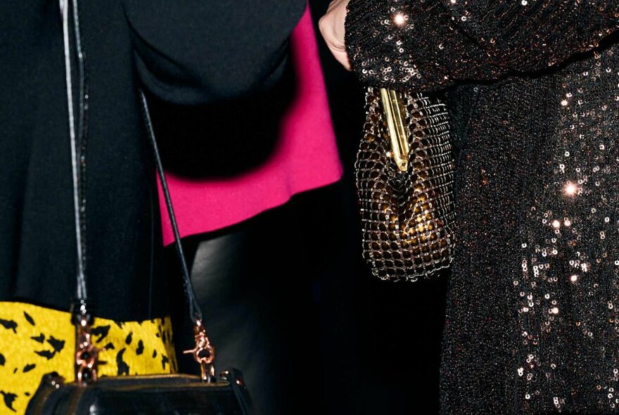 Details of two people from behind, one clutching a glomesh handbag and wearing a sequined jacket, the other in a black jacket with a small black handbag over their shoulder.