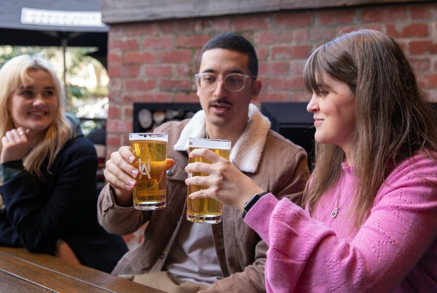 People clinking beer glasses at an open-air table in front of a brick wall.