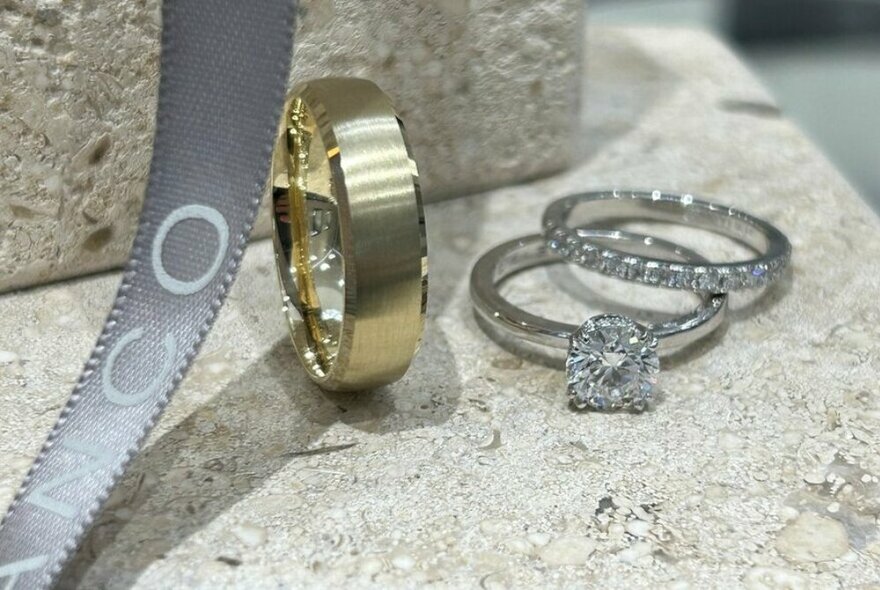 Two diamond rings displayed on a marble counter and a plain gold wedding band.