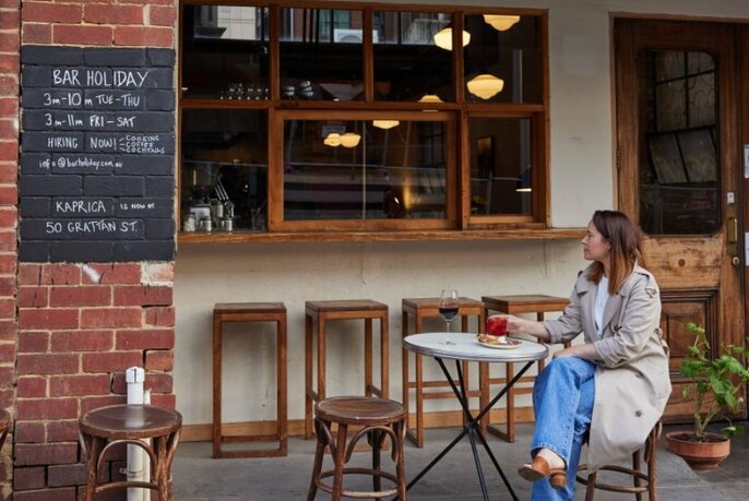Woman in trench coat on outdoor table looking up at chalkboard menu on brick wall and wooden window service nook of Bar Holiday.