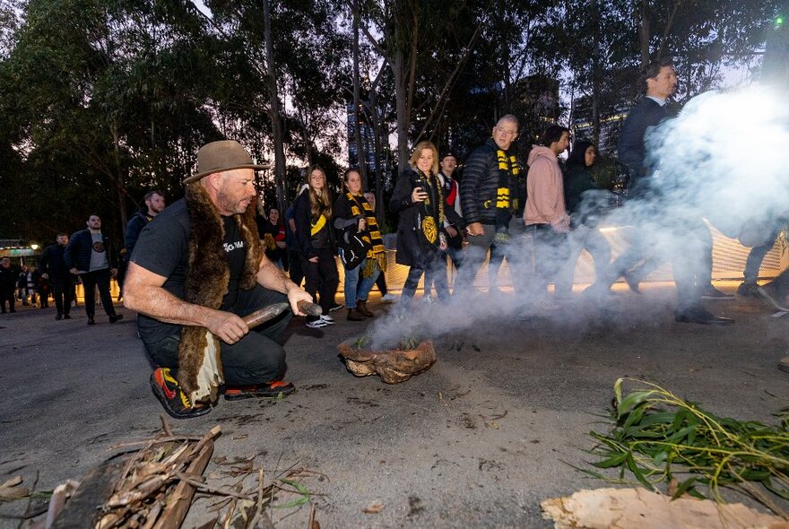 A traditional First Nations smoking ceremony with an Indigenous man wearing a traditional animal skin, and Richmond supporters walking behind.