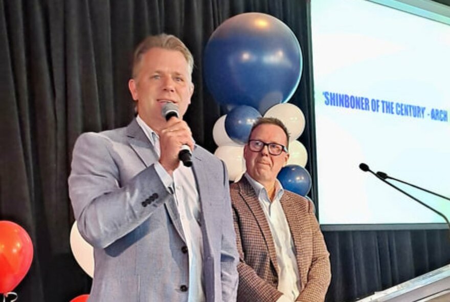 Two men wearing suits standing on a stage giving a talk, one of them speaking into a microphone, balloons and a large screen behind them.