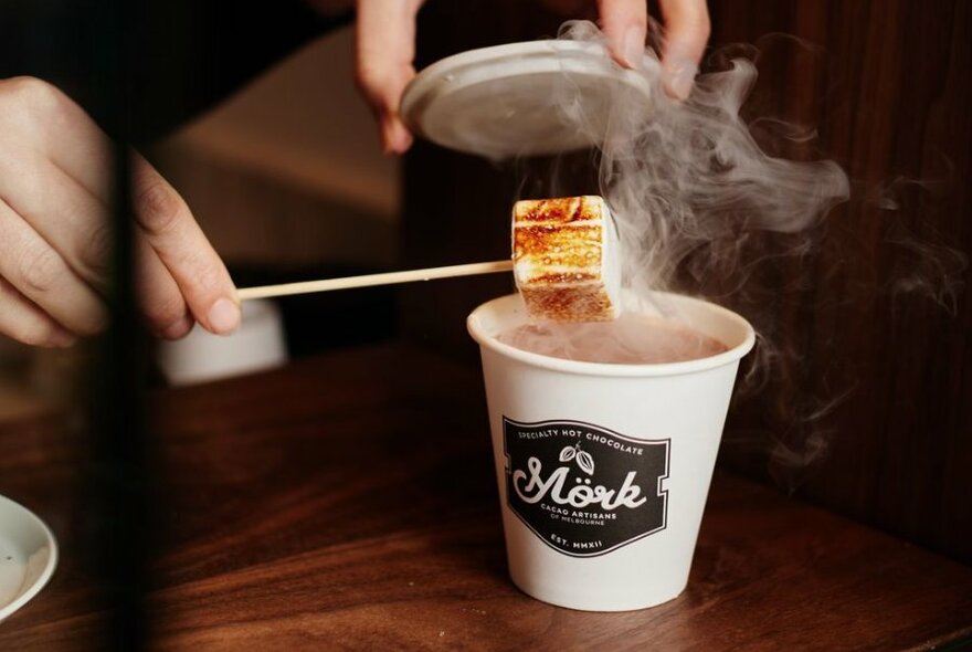A woman putting a marshmallow into a steaming takeaway cup of hot chocolate.