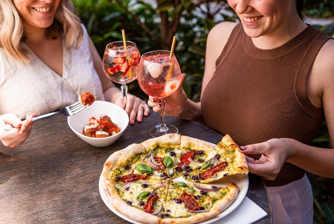 Two people at an outdoor dining table eating pizza and pasta with drinks also on the table.