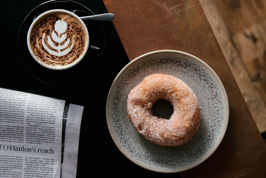 Table with a cup of coffee, donut on a plate and a newspaper.