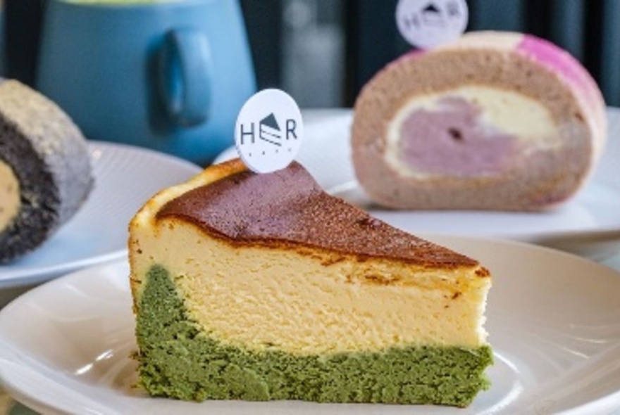 A slice of cheesecake showing a green layer at the bottom and a shop tag in the top with other cakes on plates behind. 