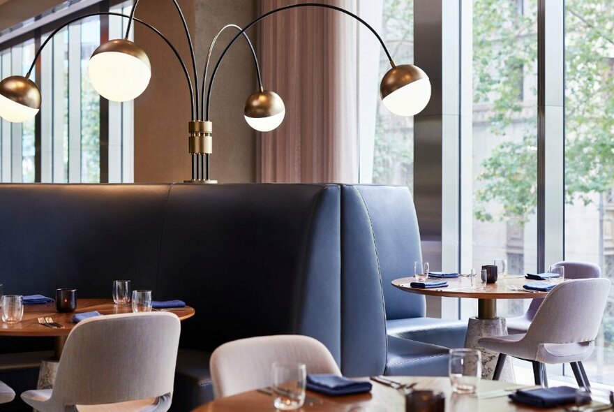 Interior of a restaurant showing comfortable  booth seating, tables and white dining chairs, with large floor to ceiling windows.
