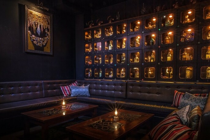 Dimly-lit corner of a bar with long, lounge-style seating and lights in small wall niches. 