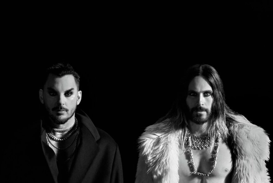 A black and white photo of two band members from the band, Thirty Seconds to Mars, one with short hair, wearing black, and the other with long hair, wearing an open fur coat with chunky jewellery; both with black eye makeup.