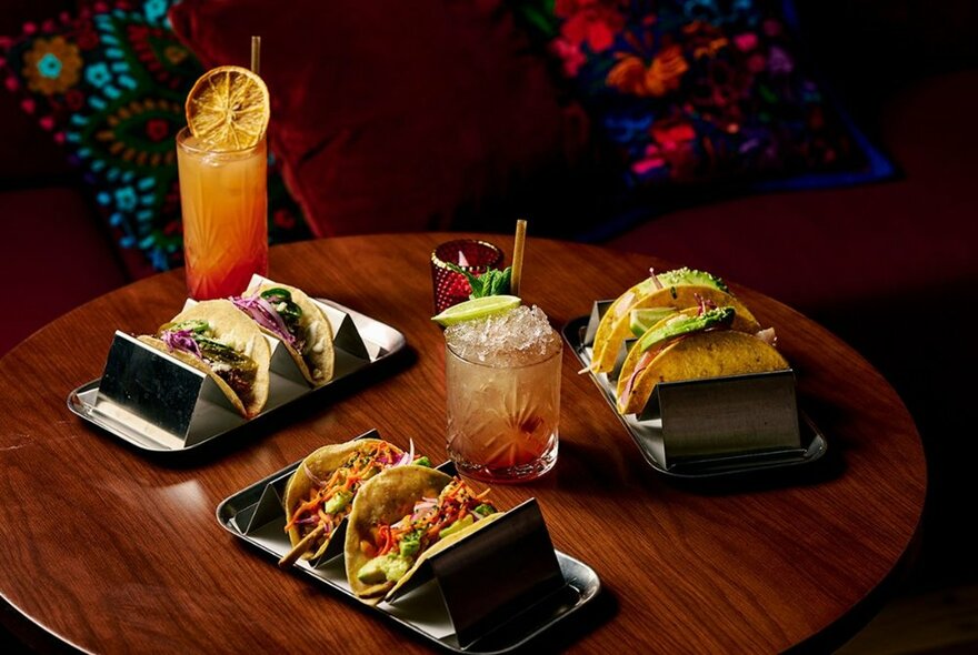 A table set with three different plates of tacos and two cocktails. 