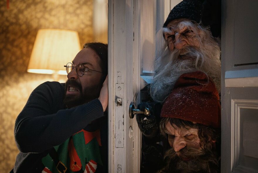 Two creatures with craggy scary faces and long white beards trying to get into a room by pushing against a door that a person is braced against on the other side in an effort to keep it closed.