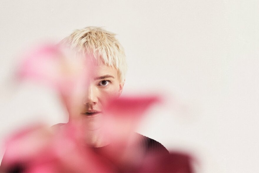 Person with short blond cropped hair, looking directly at the viewer, their face half obscured by an out-of-focus pink flower in the foreground. 