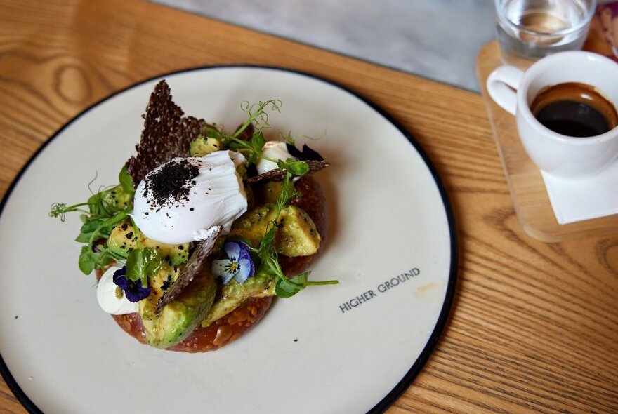 Short black espresso and a white plate with avocado toast topped with a poached egg on a wooden table.