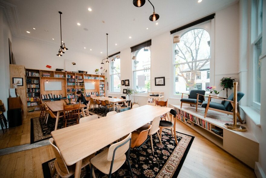 View of a bright and airy writing studio space, with walls of bookshelves, white walls and large windows, tables and writing benches, chairs and rugs.