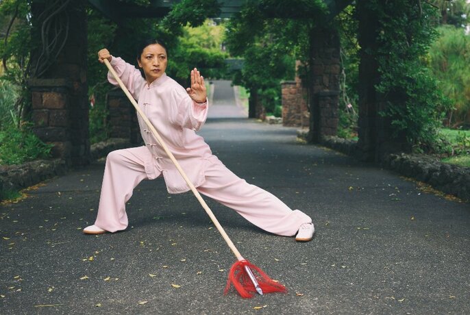 Person in a loose fitting shirt and pants, standing  in a tai chi style pose and holding a long pole, on a path in a park.