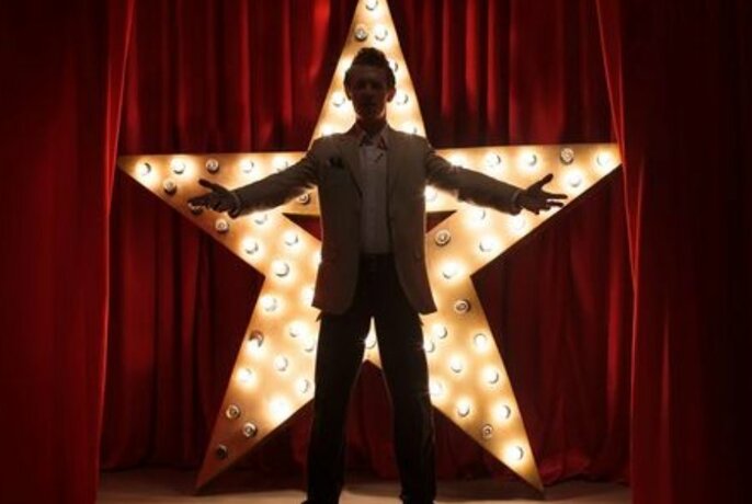 Person in silhouette with outstretched arms, standing in front of an illuminated star on a stage, with red velvet curtains behind them.