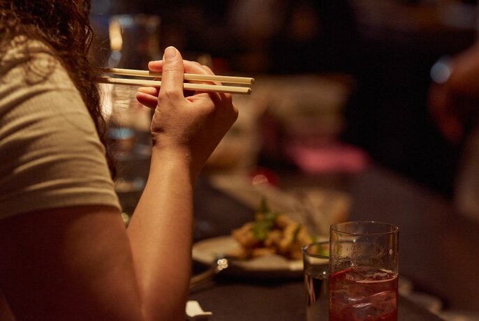 Hand holding chopsticks with dishes of food on a table in the background.
