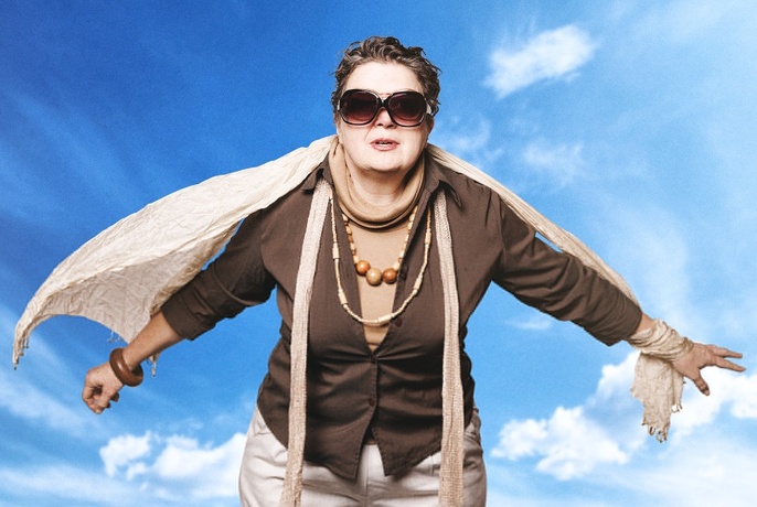 Older woman wearing brown and beige, posed as if flying, with blue sky behind.