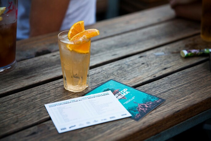 Cocktail in a tall glass, garnished with slices of orange, resting on a wooden table next to a Rooftop Cinema program flyer.