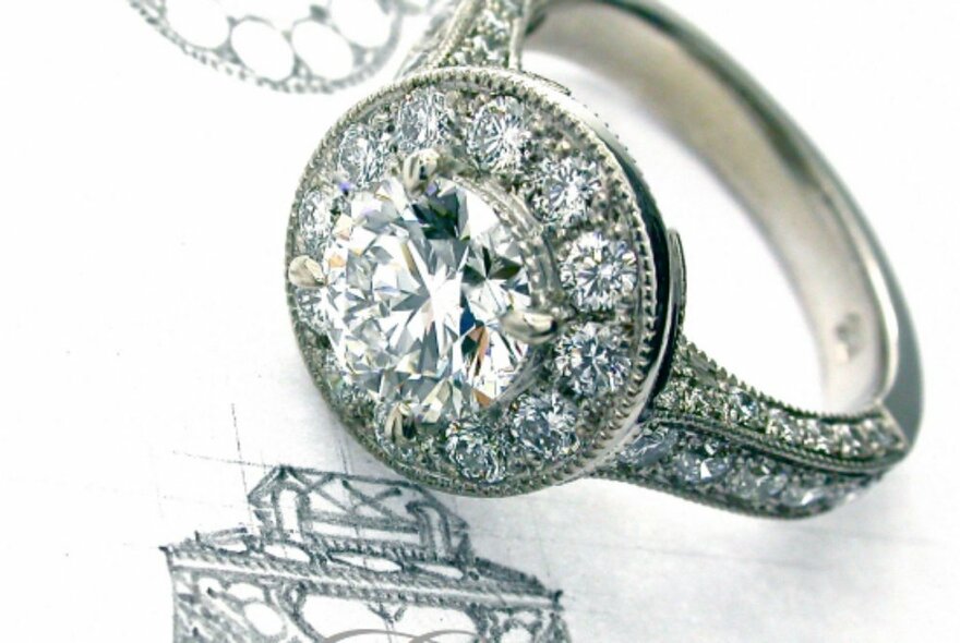 A diamond encrusted white gold engagement ring with a round cut diamond in the centre, resting on a paper with its matching hand-drawn design.