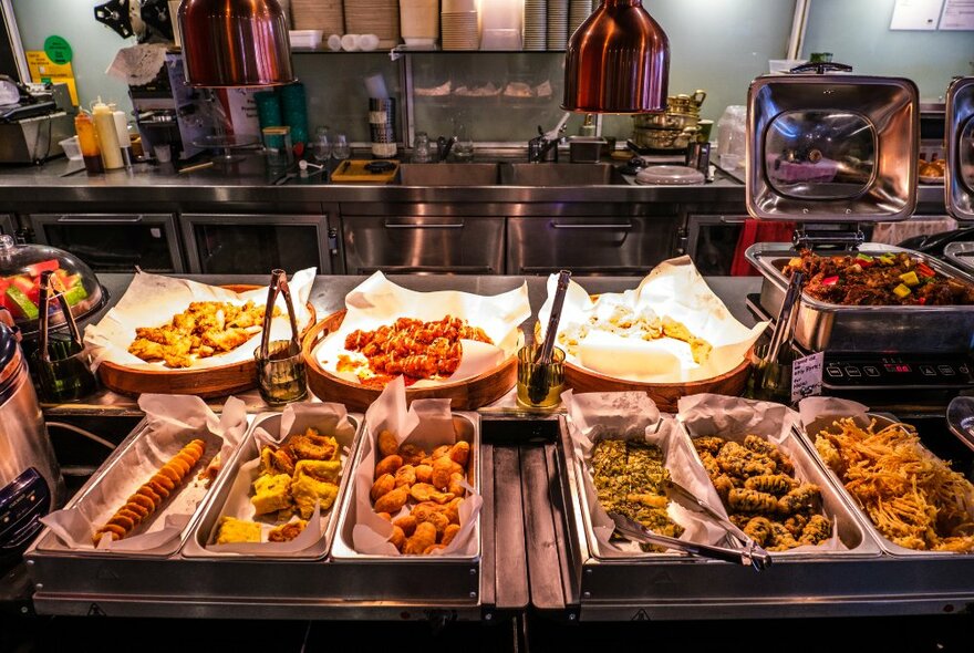 Dishes of hot food in trays on a restaurant kitchen counter, with cooking equipment in the background.