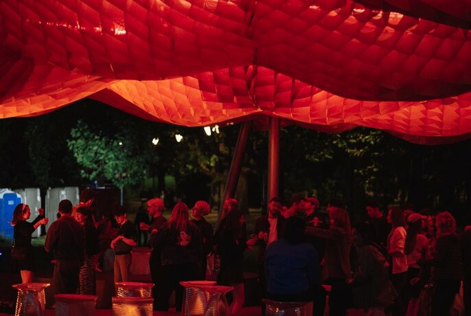 The MPavilion building at night, featuring a red-lit sail design, in a park with people gathering around.