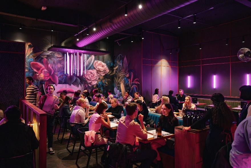 Interior of a restaurant illuminated with soft pink wall lights, patrons eating and drinking at tables, a large floral mural on the read wall.