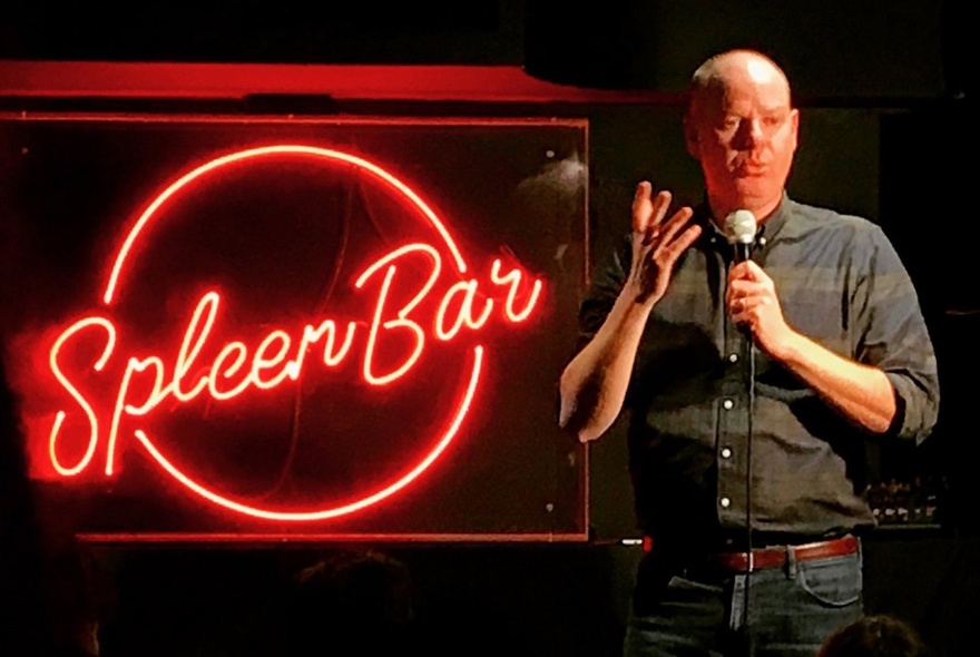 Stand-up comedian Tom Gleeson on stage in front of Spleen Bar neon sign, holding microphone and telling a joke.