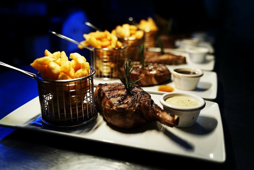 Row of platters with chips in baskets, steak and bowl of sauce.