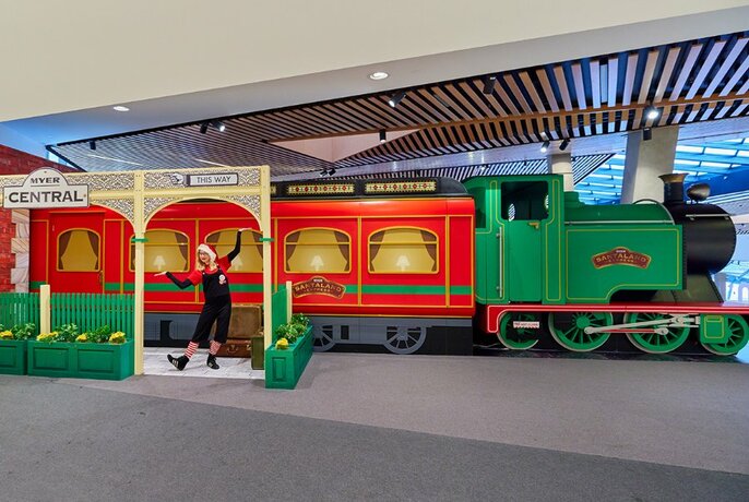 A Christmas helper poses in front of a large red and green train.