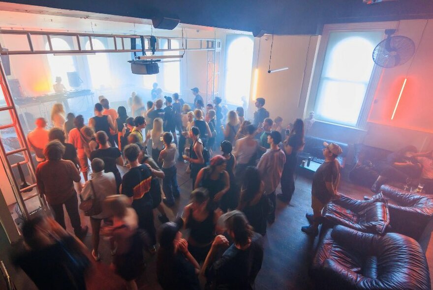 Overhead view looking down into a large room with a small group of people dancing in front of a DJ who is playing at one end of the room; there are large armchairs at the other end of the space and the windows have light streaming in. 