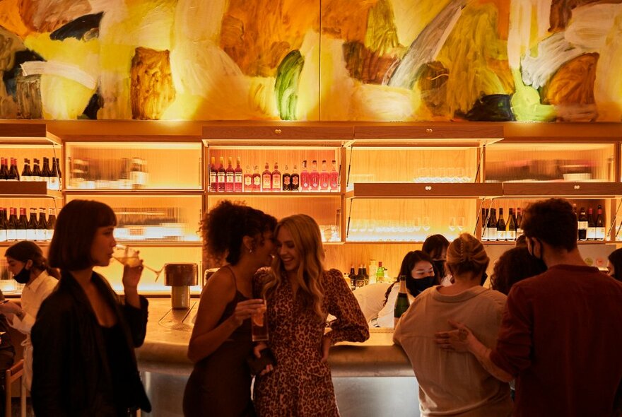 People leaning against a bar, sipping drinks and close talking, the bar area and bottles on shelves behind the bar bathed in a warm orange-yellow light.
