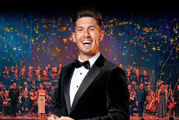 Man wearing a tuxedo, smiling in front of a large orchestra and performers on stage.