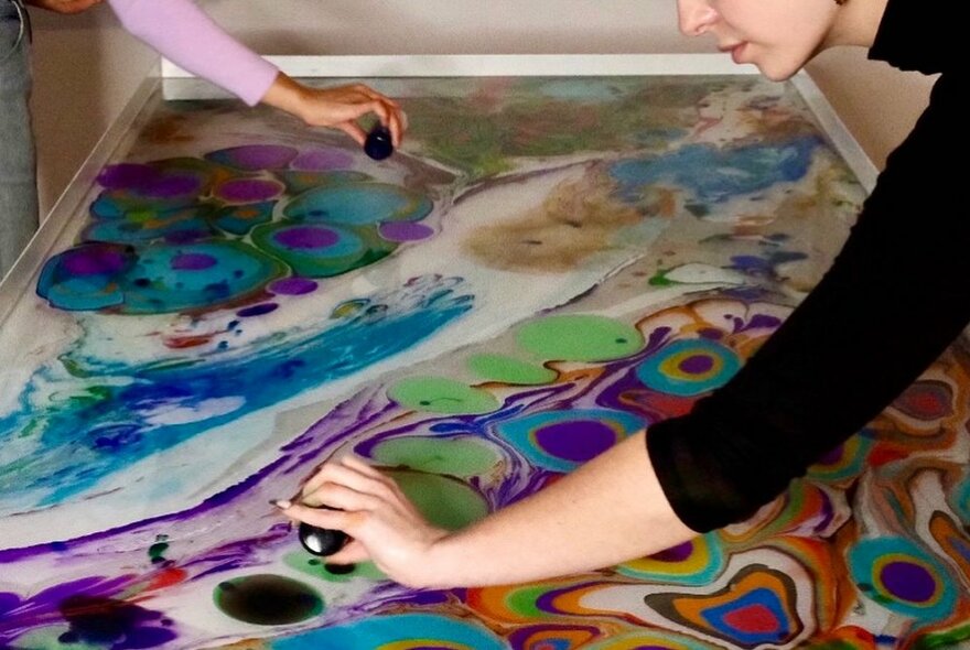 Hands pouring ink from small bottles onto a screen print of a colourful ink marbled pattern.