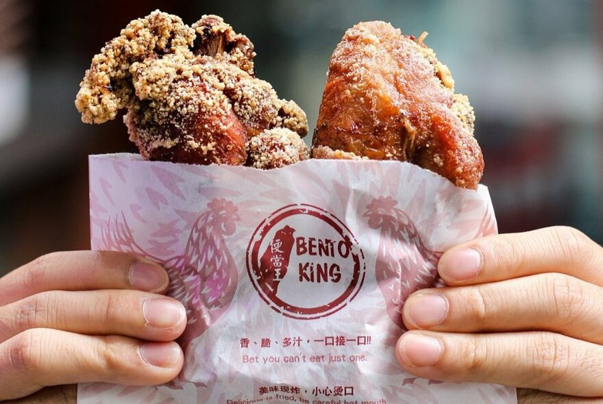 Pair of hands holding up crispy fried chicken in a paper bag with the word Bento King written on the outside of the bag.